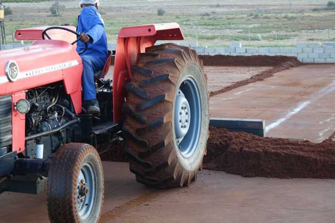 Ein Traktor mit einem 4 Meter breiten, quer montierten Brett am Heck schiebt den fermentierten Rooibos vom Rand in die Mitte des Tea Courts. Während ein Teil bereits verschoben wurde, ist im Hintergrund die noch angehäufelte Rooibos-Reihe am äußeren Rand des Tea Courts zu sehen, die ebenfalls noch in die Mitte geschoben werden wird.