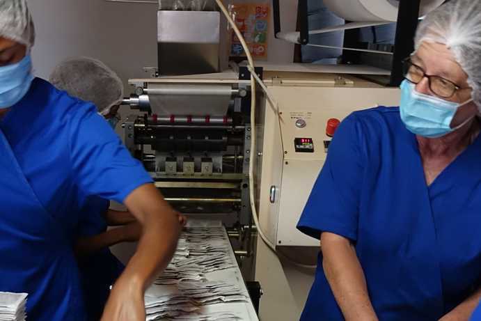 The image shows the tea bag manufacturing machine on Klipfontein Farm from the front view. In the lower part of the machine, the finished, sealed Rooibos tea bags are being ejected. Above, it is clearly visible how the filter material is fed into the machine from a roll before entering the production process.