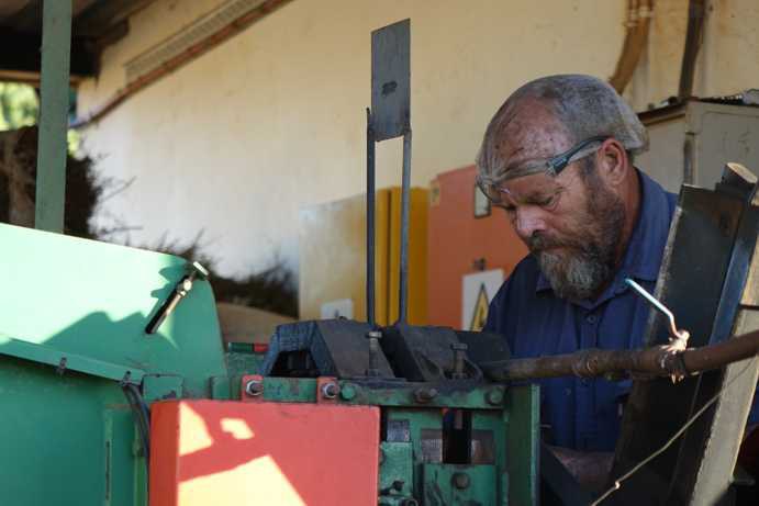 A machine operator inspects the cutting system for Rooibos processing. He adjusts the guillotine cutter, which consists of four star-shaped, 90° offset cutting blades. These rotate at high speed to chop the Rooibos branches. Due to heavy wear, the blades must be resharpened hourly using a grinding tool.