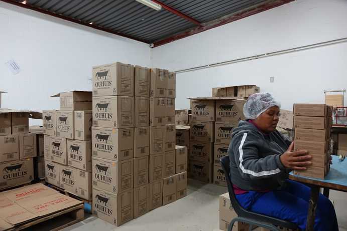 Une travailleuse emballe du thé Rooibos Ouhuis dans des cartons de conditionnement plus grands. Plusieurs palettes remplies de cartons d’expédition Ouhuis sont empilées dans la pièce. La vente directe sous sa propre marque permet de préserver les emplois locaux sur la ferme.